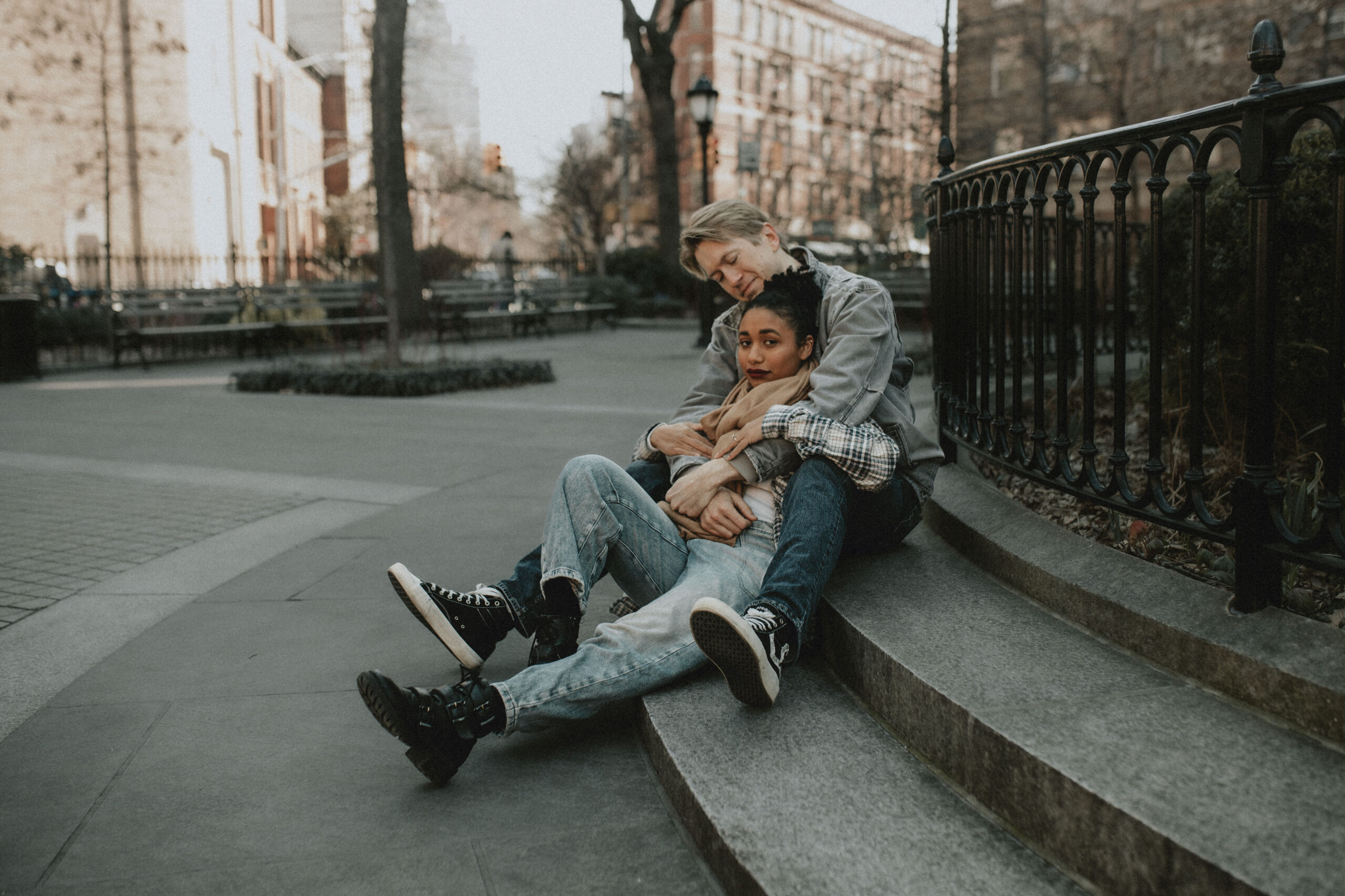 A couple in 1990's retro vibe clothing sits in a little park in Manhattan, cuddled together on the ground during their engagement session