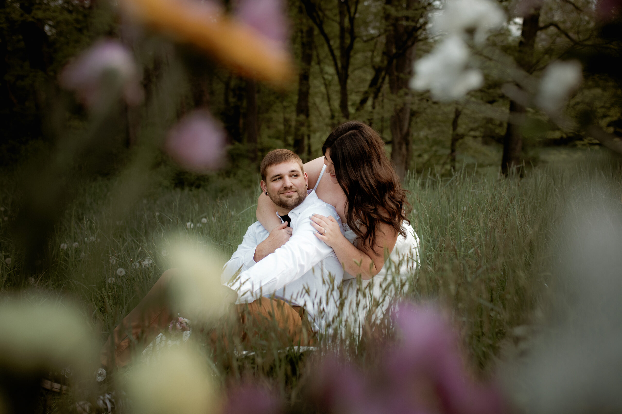 A woman hugs a man from behind while kneeling on a blanket for their springtime Connecticut engagement photos - while framed in colorful wildflowers