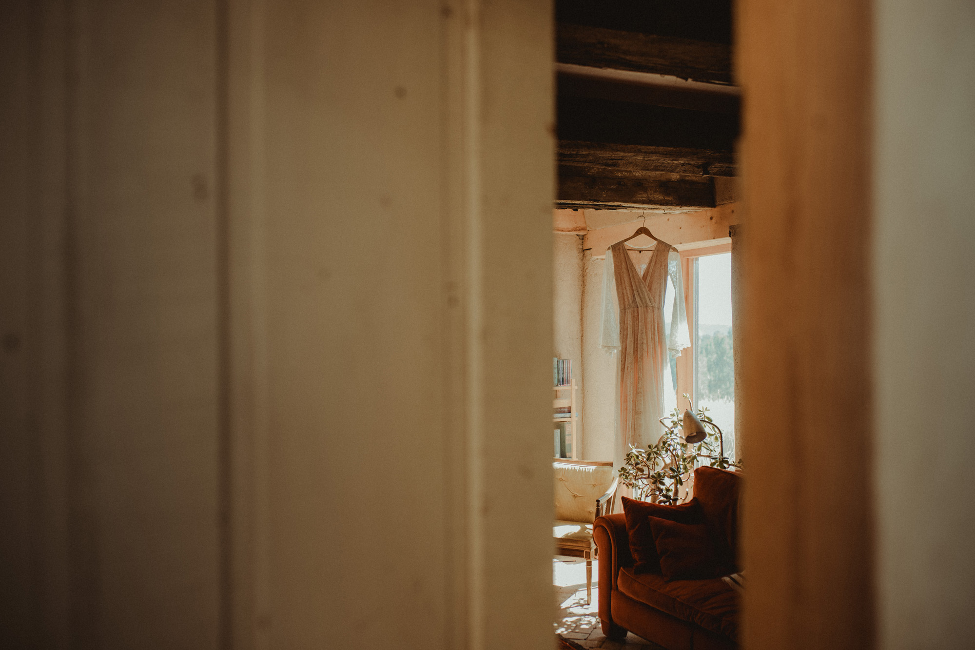 A wedding dress hangs framed in front of a window, as seen through a small crack in the door to the room