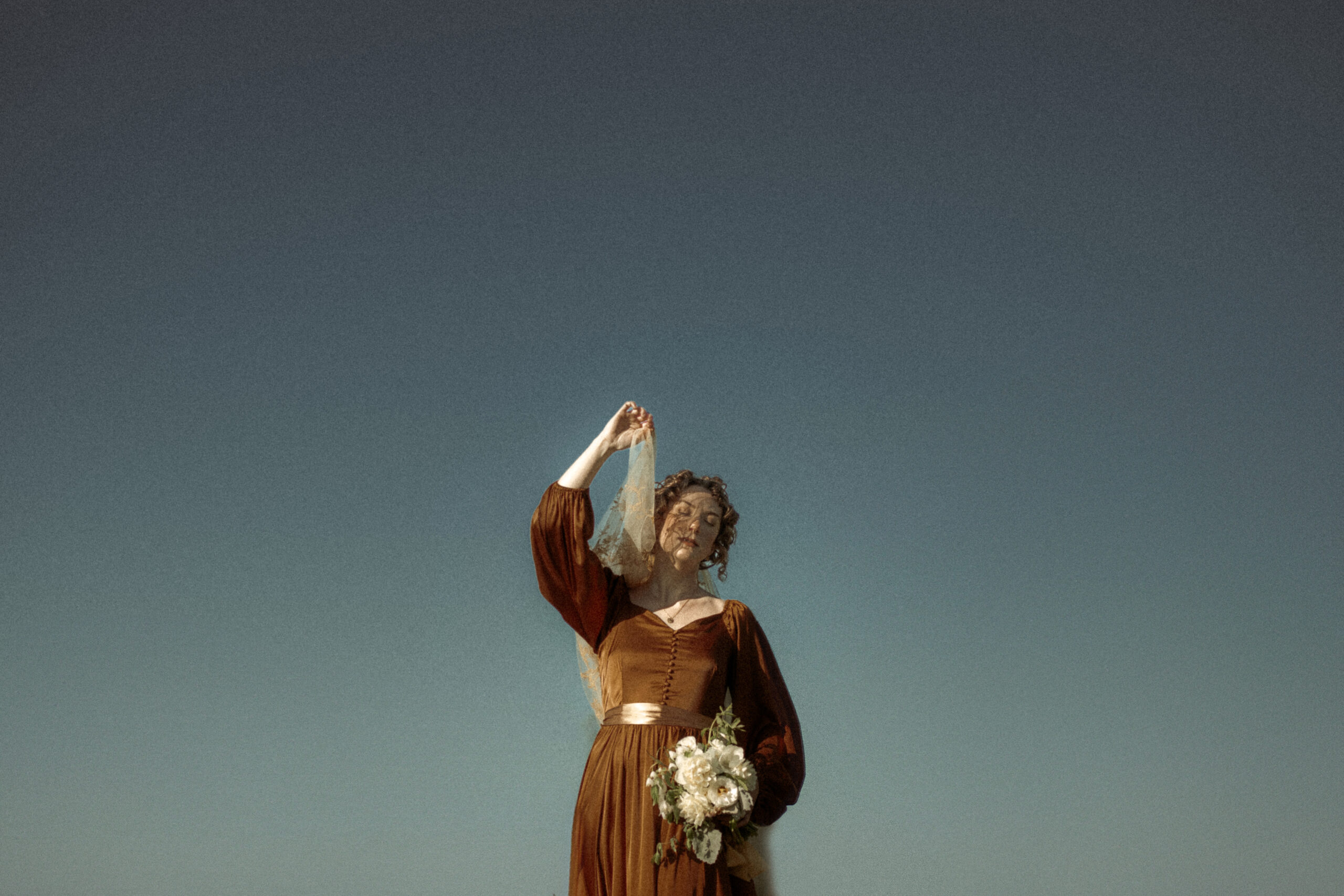 A bride in a gold satin dress with a gold embroidered veil stands against a vivid blue sky