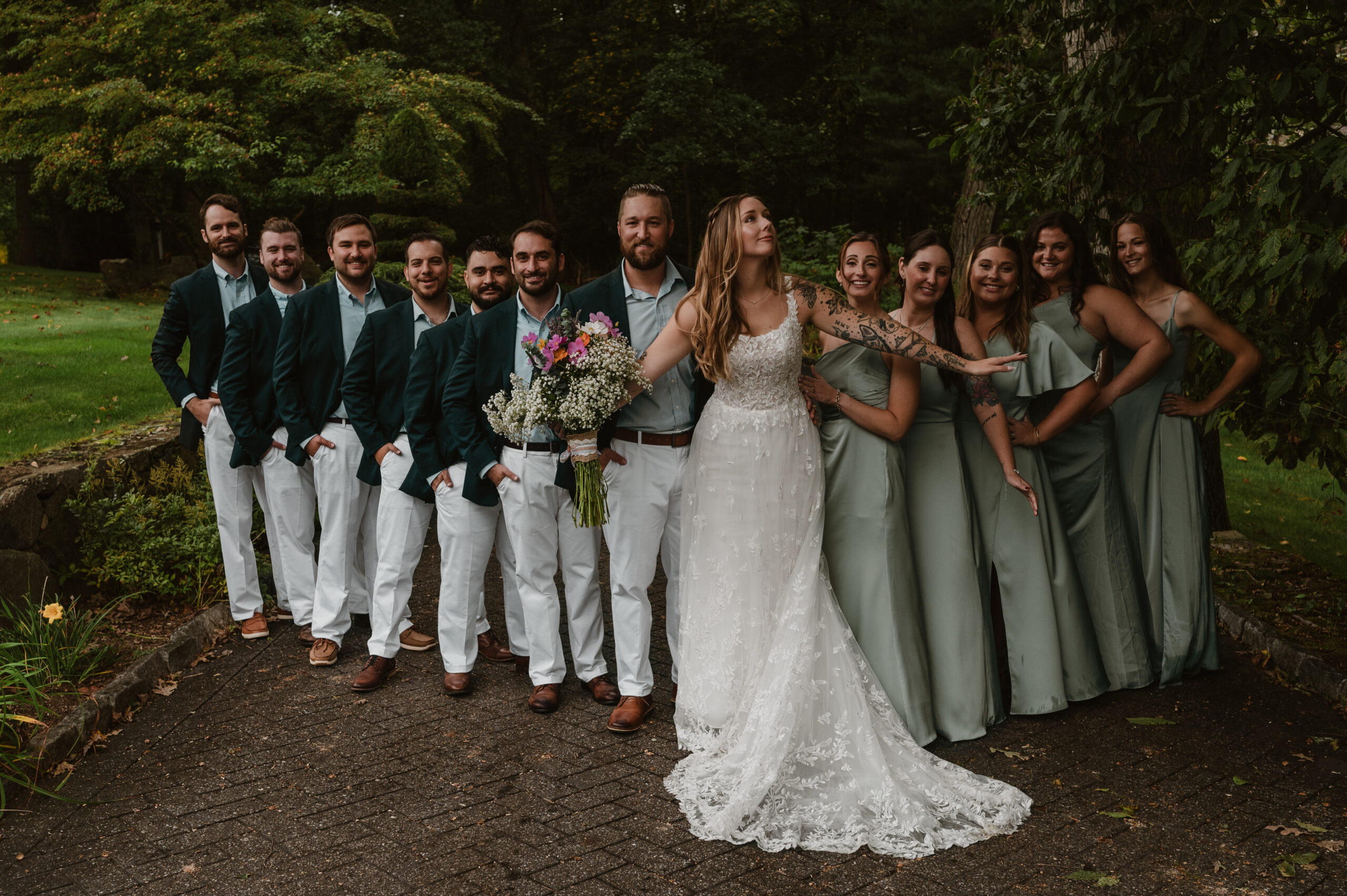 A bride stands in front of her groom and wedding party with arms spread wide on her rainy micro-wedding day