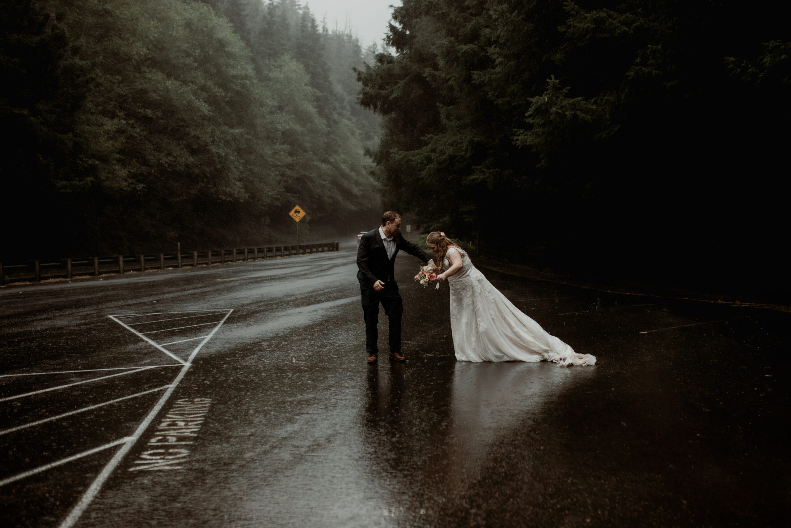 a bride and groom stand in an empty parking lot in the pouring rain, surrounded by mist and dark trees