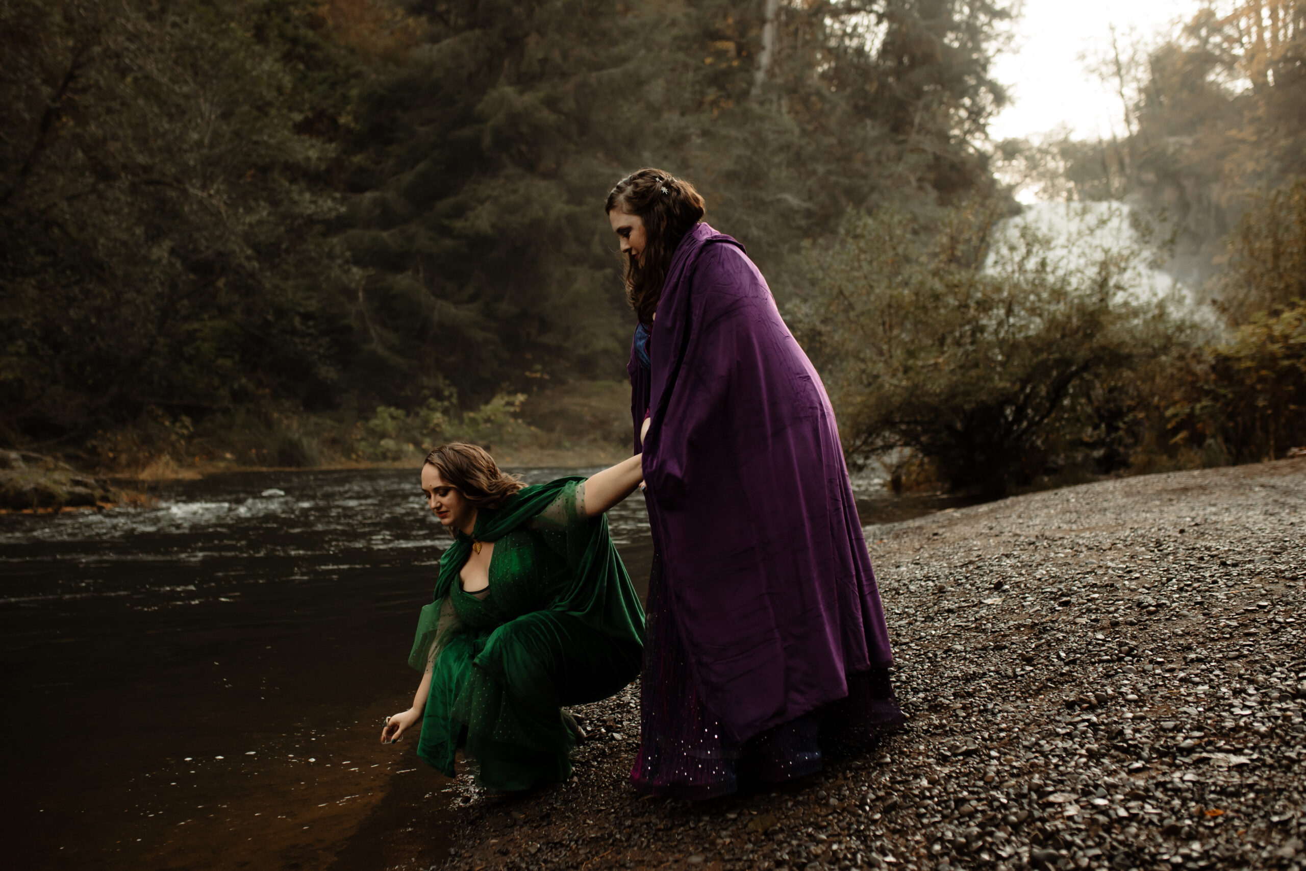 two brides, one wearing emerald green and the other wearing royal purple, stand next to a river in Oregon, one bending down to collect water in a little jar for their wedding day keepsake